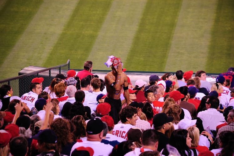 Sports fans in stadium watching the game while another fan, dressed I. Mock face paint and headdress, stands and does a stereotypical Indian yell by patting his hand to his mouth.